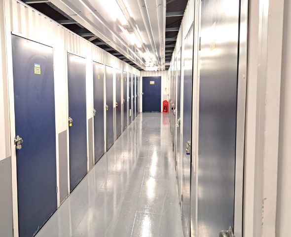 Interior view of the Safestore self-storage facility in Milton Keynes showing a clean, well-lit corridor with multiple blue storage units on either side. The shiny epoxy-coated floor reflects the overhead fluorescent lighting, and each door is number