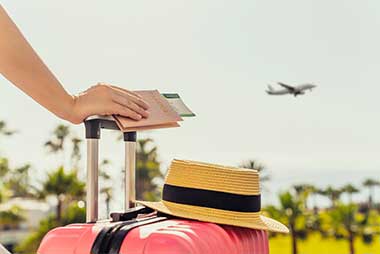 Close-up of a person resting a hand on a pink suitcase with a blue sky and airplane in the background, suggesting travel