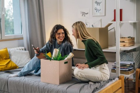 Two female students sitting on a bed in a university hall room, smiling and packing belongings into a storage box