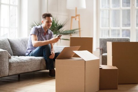 Packed cardboard boxes in a living room with a person on the sofa using a phone while preparing to move home