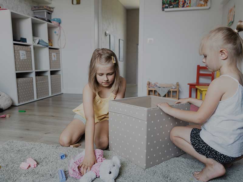 Two children playing in a cluttered room with toys and storage bins, illustrating summer holiday chaos and the need for home organisation.