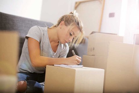 Person sitting on a couch writing in a notebook on top of a cardboard box, surrounded by moving boxes, suggesting preparation for a summer move.