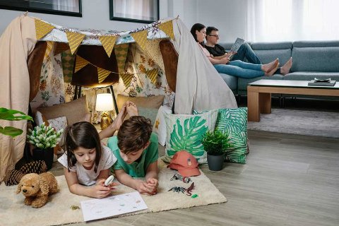Children drawing inside a blanket tent in a family living room during summer holidays, surrounded by toys and bunting