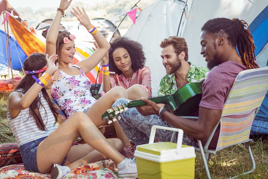 Group of friends sitting on the ground at a campsite with tents in the background, one person playing a guitar while others raise their hands and enjoy the music.