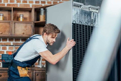 Man in overalls carefully moving a fridge freezer, ensuring safe transport in a kitchen with wooden shelves in the background.