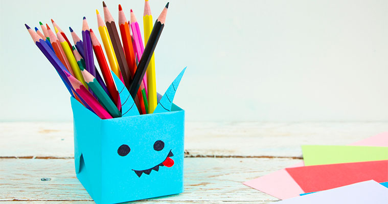 A blue pencil pot with a face on it made out of cardboard filled with colouring pencils