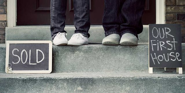 Two people standing on a doorstep with 'Sold' and 'Our First House' signs, representing new beginnings and shared spaces