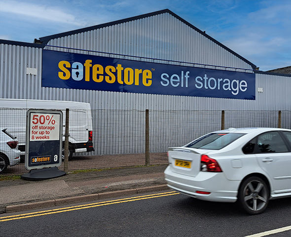 Exterior view of the Safestore self-storage building in Milton Keynes. The large facade features the company's branding in blue and yellow against a grey corrugated metal structure. A fence is visible in the foreground, and a promotion banner.