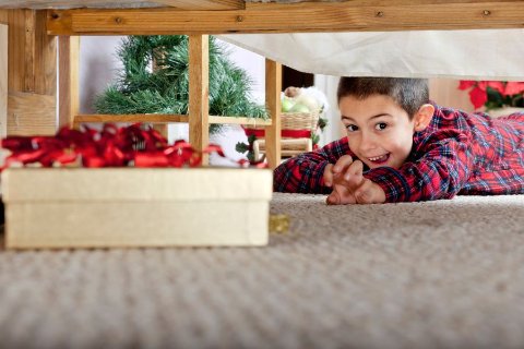 Child in Christmas pyjamas looking under the bed and finding a hidden present beside a Christmas tree