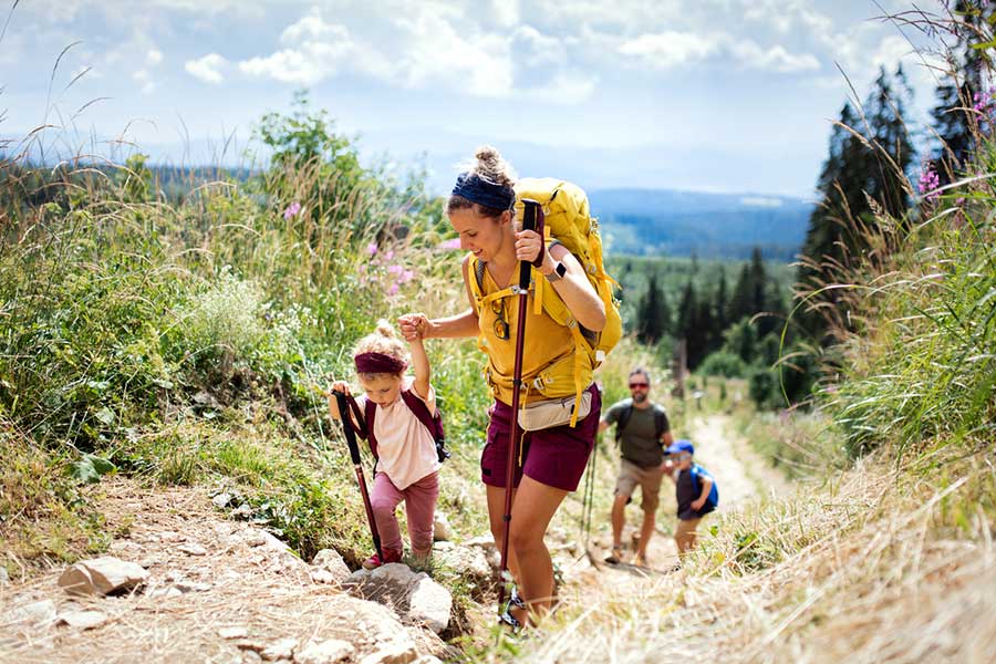 Family hiking up a rocky mountain trail with lush greenery and distant peaks.