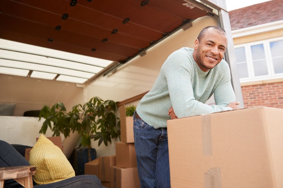 Smiling man leaning on a moving box beside a van loaded with boxes, furniture and a plant