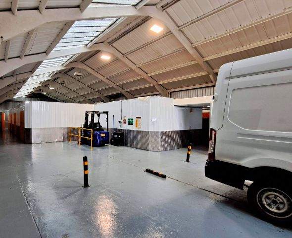 Image of a loading bay at Safestore self-storage facility in Milton Keynes. The bay is indoors with a high ceiling, featuring a white delivery van parked on the right and a blue forklift in the center. 