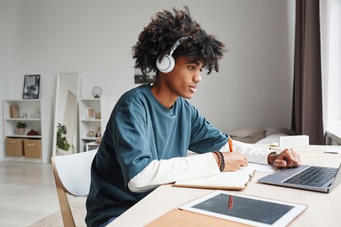 Student wearing headphones studying at a tidy desk with a laptop, notebook, and tablet in a bright, organised room.