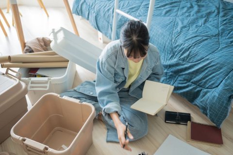 A student sitting on her bedroom floor surrounded by storage boxes, making notes while packing up her belongings at the end of term